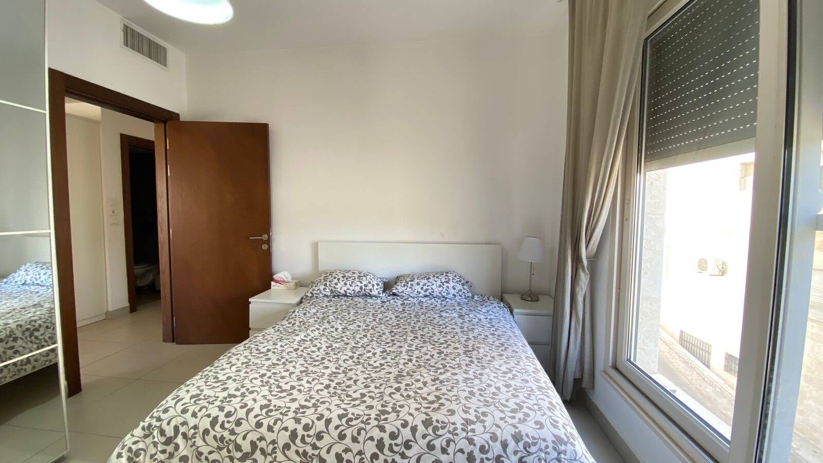 b4083283-11d8-4f21-b9a3-1d6e2cd8a6fe Bright modern bedroom with a double bed, white headboard, two white nightstands, and a lamp on the right; large window with beige curtains on the right side fills the room with light.