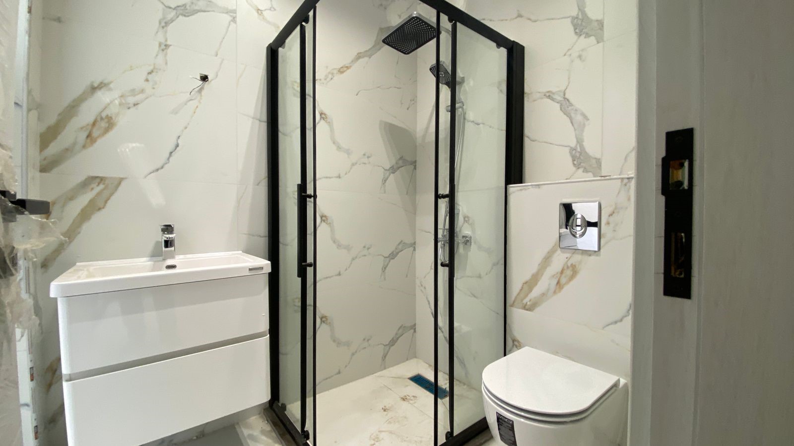 b425fc01-3afa-4776-9a18-1078e7ee2a72 Modern bathroom with a glass-framed corner shower, white vanity, and wall-mounted toilet set against marble walls.