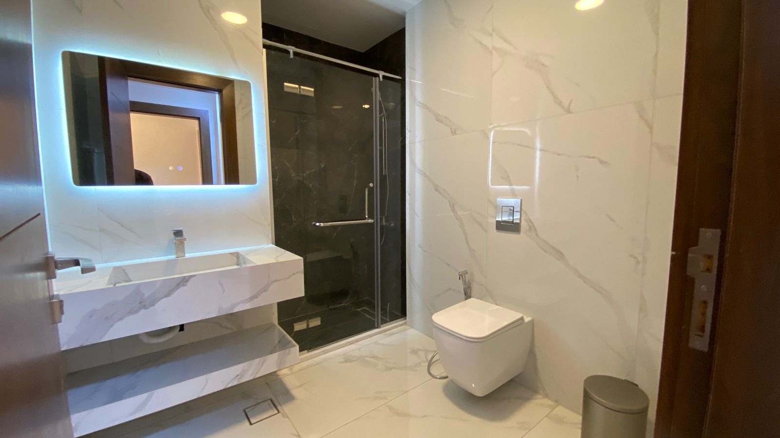 b4320282-0f91-48fc-9b60-a1bcc5ecfeb5 Modern marble bathroom with a wall-mounted sink and backlit mirror, glass shower enclosure, and a wall-hung toilet across from the door.