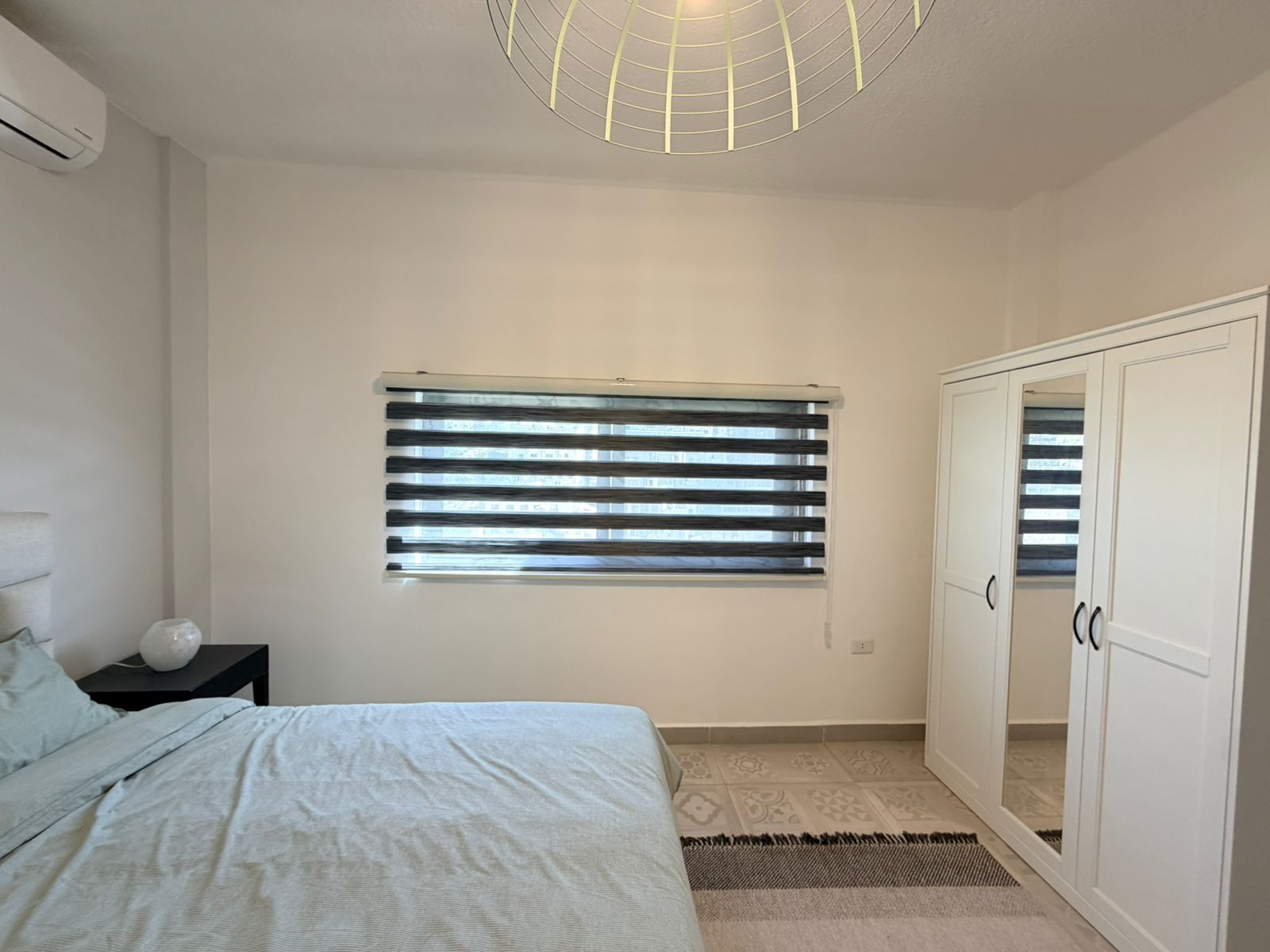 b4845932-a0a6-41a5-adce-58acceaf0356 Bedroom with a bed on the left, striped window blinds, and a white wardrobe with mirrored doors on the right.