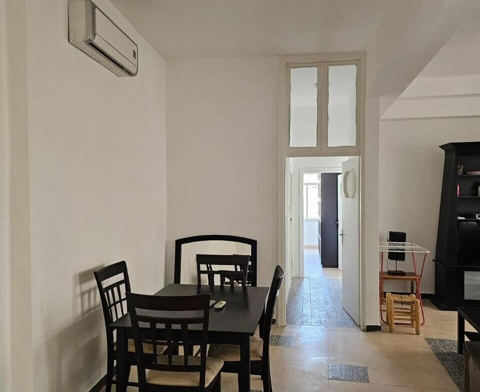b4896d52-f530-4518-87b2-234641e82f1e Dining area with a dark wooden table and four chairs against white walls; an air conditioner mounted left, doorway to a bright hallway in the back.
