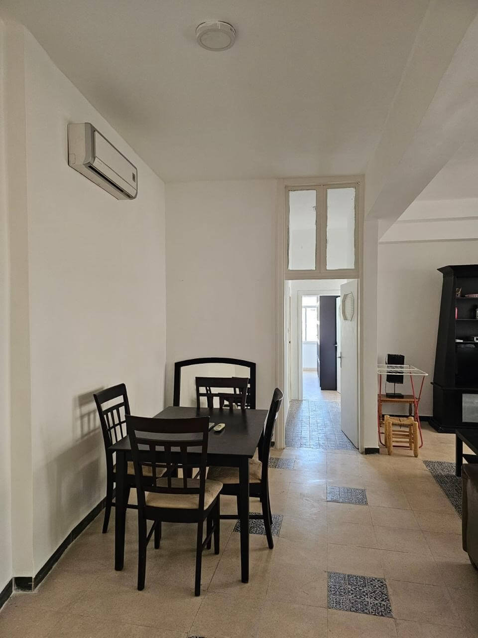b4896d52-f530-4518-87b2-234641e82f1e Dining area with a dark wooden table and four chairs against white walls; an air conditioner mounted left, doorway to a bright hallway in the back.