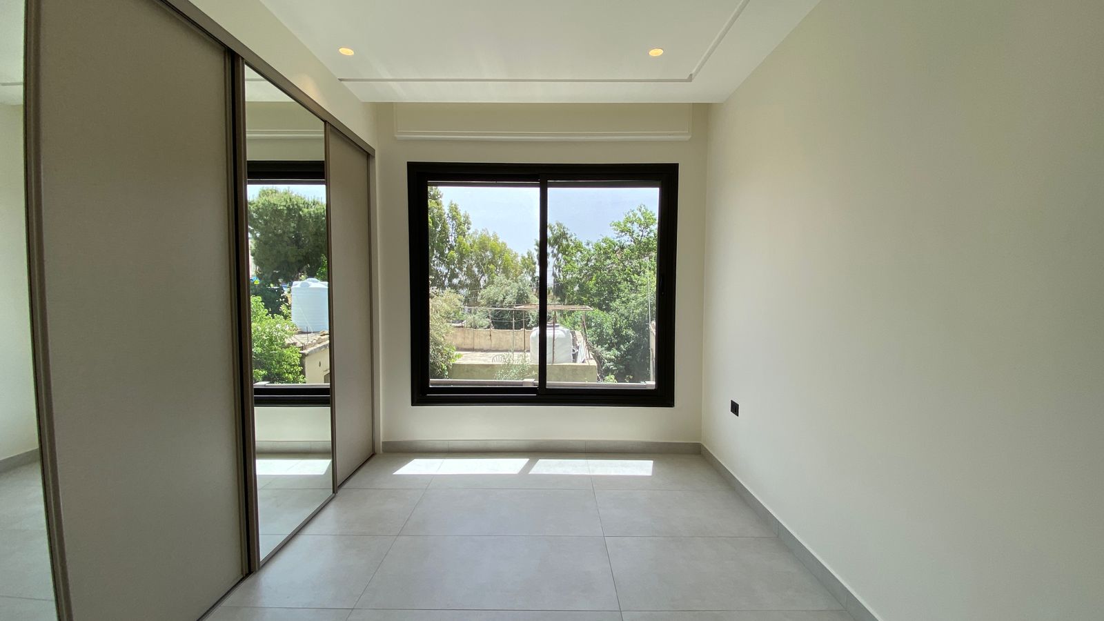 b4c25133-a6d6-46a0-8d2f-90737b4b2a89 Empty modern bedroom with large black-framed window overlooking trees; built-in mirrored wardrobes on the left.
