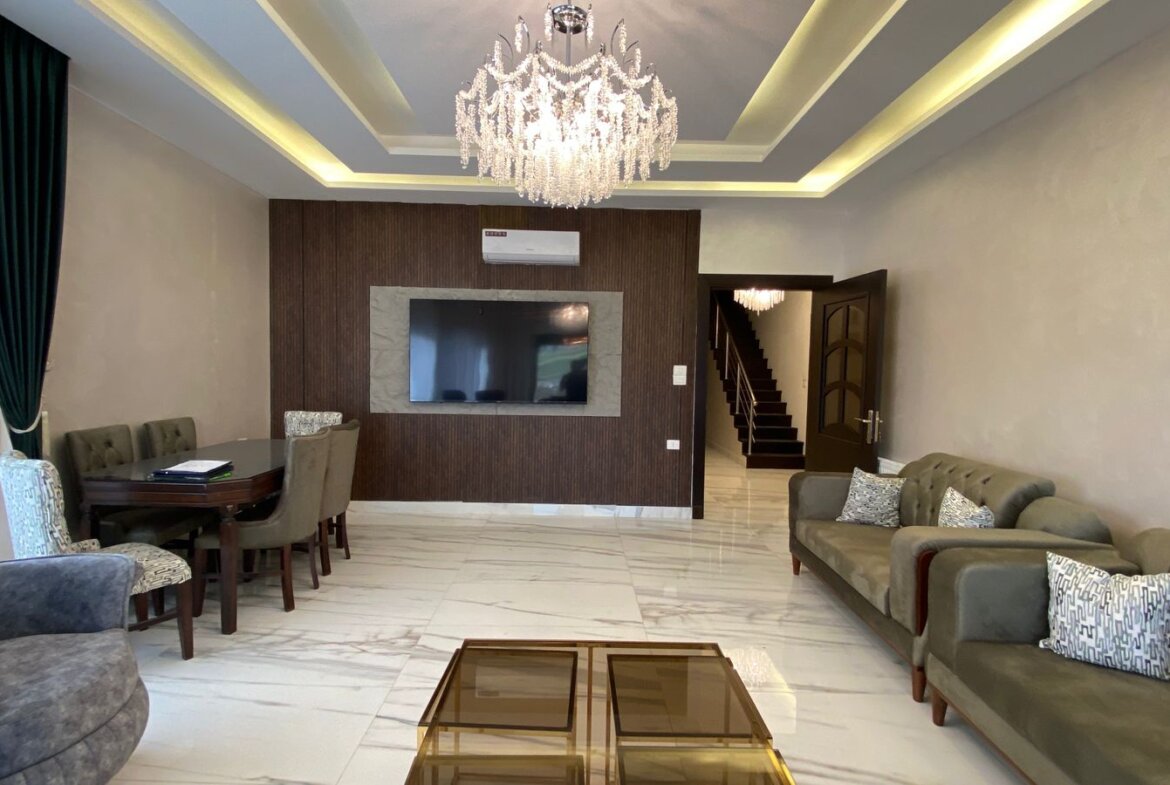 Spacious living room with a crystal chandelier, a wall-mounted TV on dark wood paneling, and light marble floors with green curtains and gray sofas.