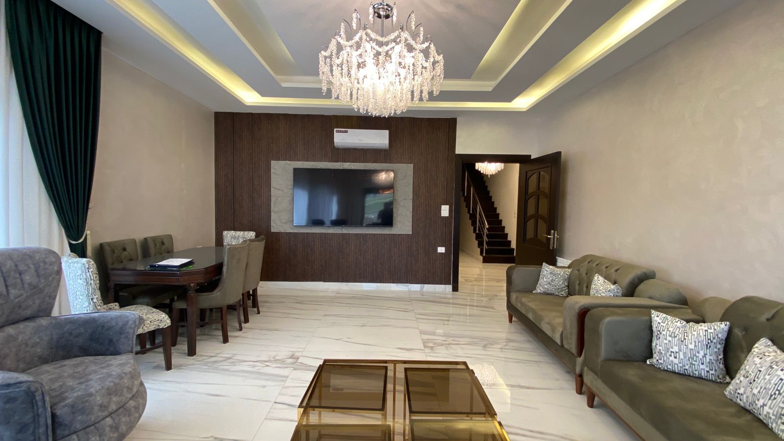 Spacious living room with a crystal chandelier, a wall-mounted TV on dark wood paneling, and light marble floors with green curtains and gray sofas.