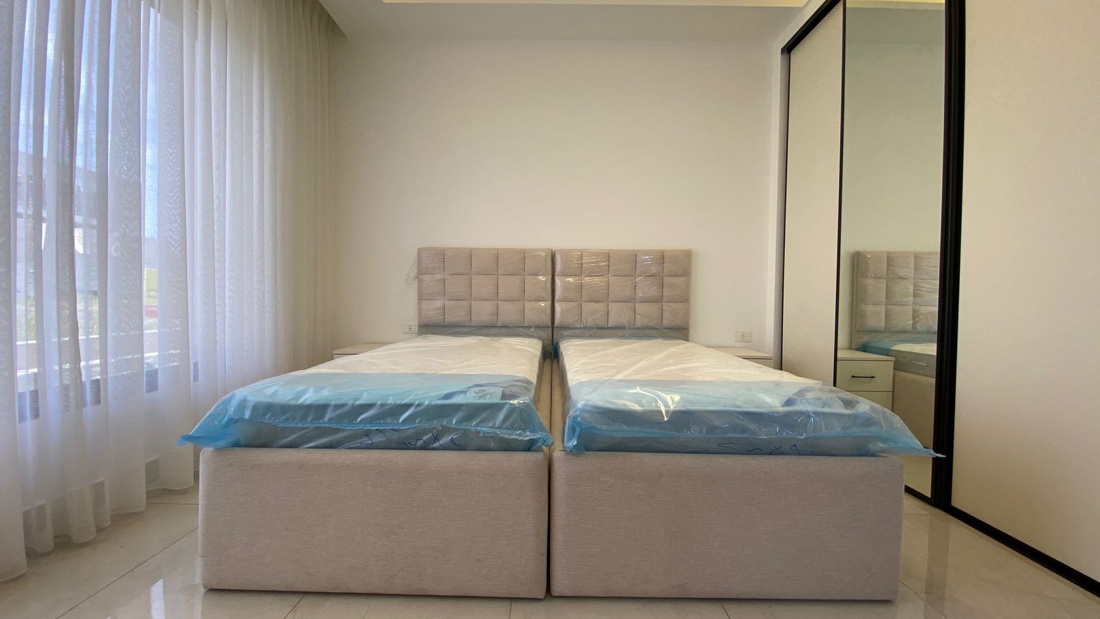 b594a730-99c0-475d-a0c4-2edd91d88b57 Two twin beds with beige upholstered headboards in a bright, minimalist bedroom, with sheer white curtains and a large mirrored wardrobe on the right.