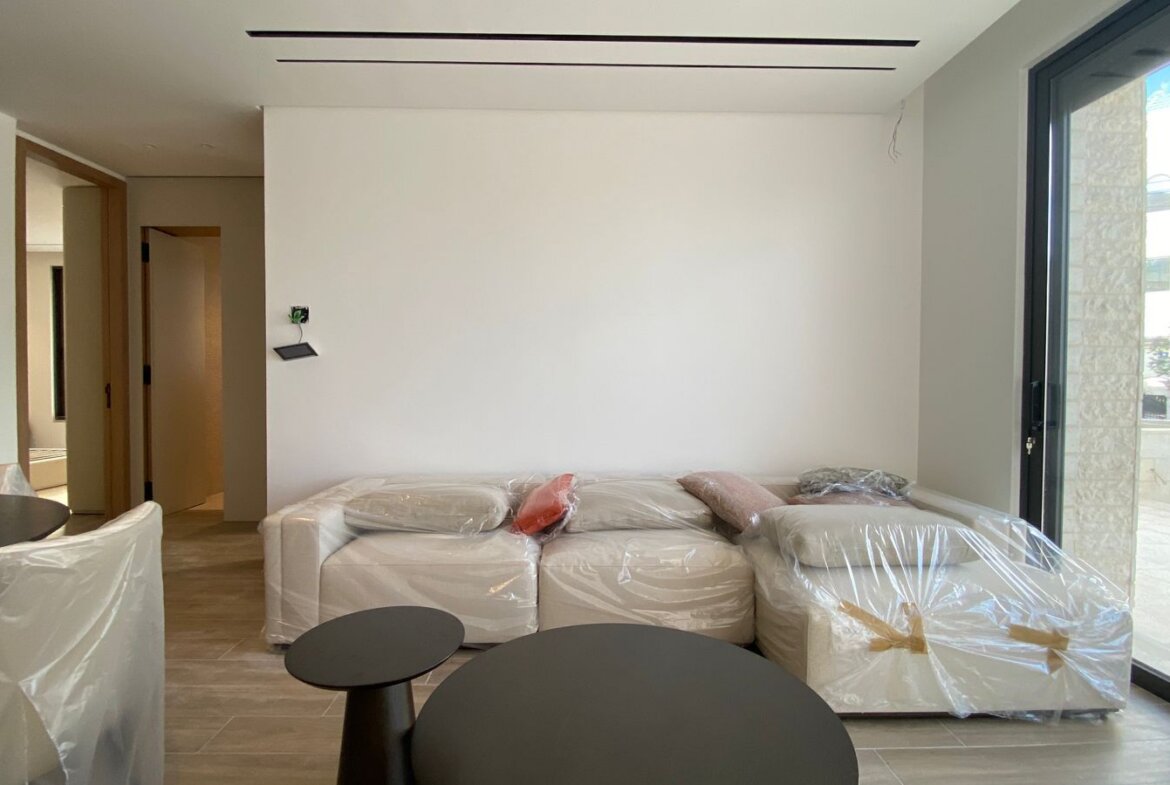 Modern living room with a white sectional sofa covered in plastic and two black round coffee tables by a large glass door.