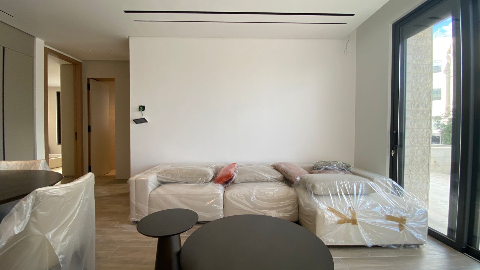 b5d1be3c-98f1-4c8d-a809-cbfd9cf324f0 Modern living room with a white sectional sofa covered in plastic and two black round coffee tables by a large glass door.