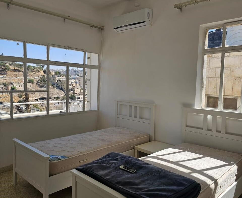 b5d66fd6-c0cc-479d-81e2-5c4b3e17d766 Bright white bedroom with two single beds, a small nightstand, and a wall-mounted air conditioner; large windows overlook a cityscape.