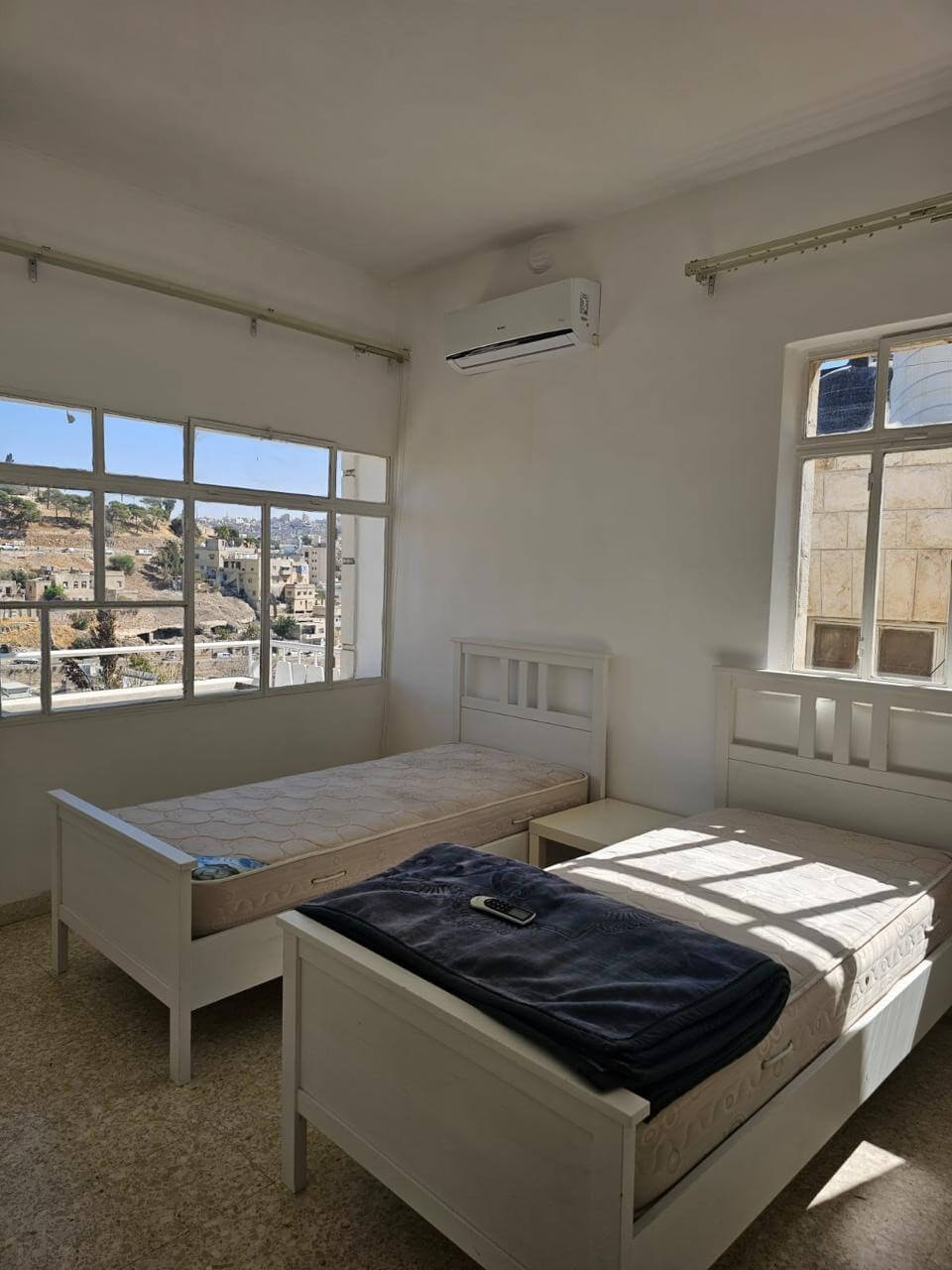 b5d66fd6-c0cc-479d-81e2-5c4b3e17d766 Bright white bedroom with two single beds, a small nightstand, and a wall-mounted air conditioner; large windows overlook a cityscape.