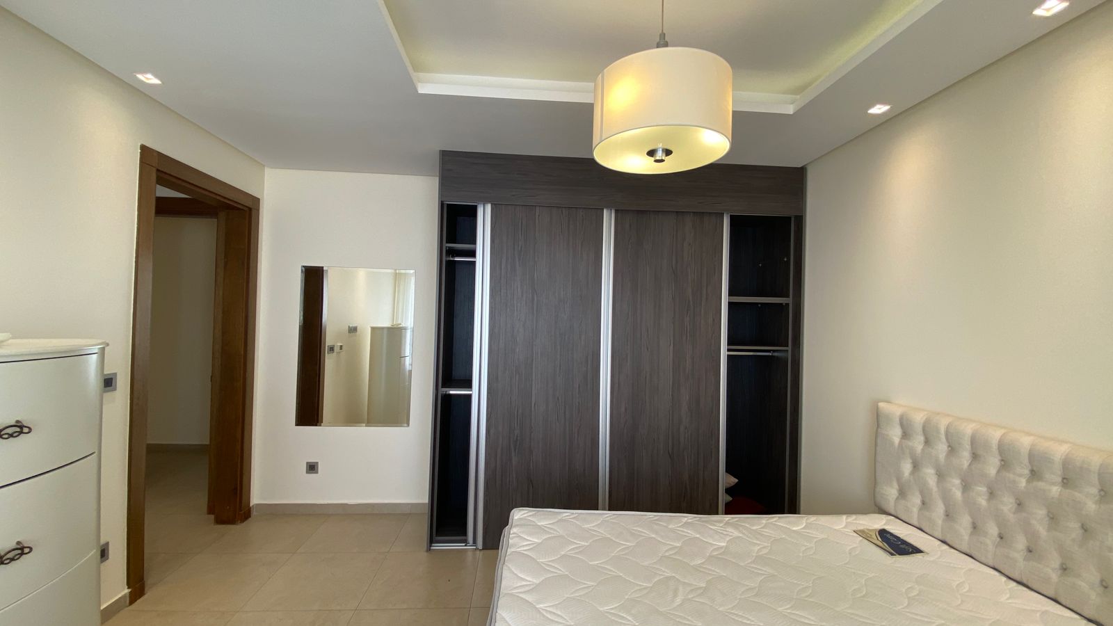 b636d6e7-8a20-4b1a-907b-15f39c63422e Modern bedroom with a large dark wood sliding-door wardrobe and a tufted upholstered headboard bed, mirror on the wall, and a white dresser to the left.