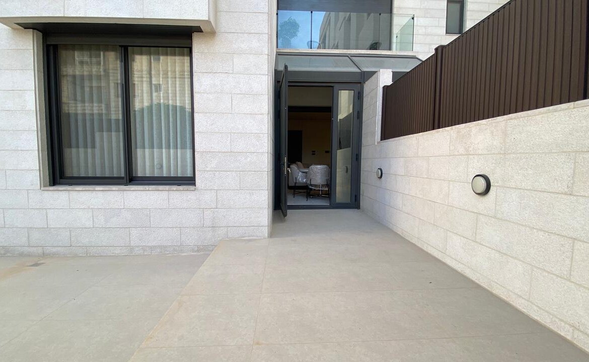 Open glass door into a modern apartment; beige stone walls, outdoor patio, and seating visible inside.