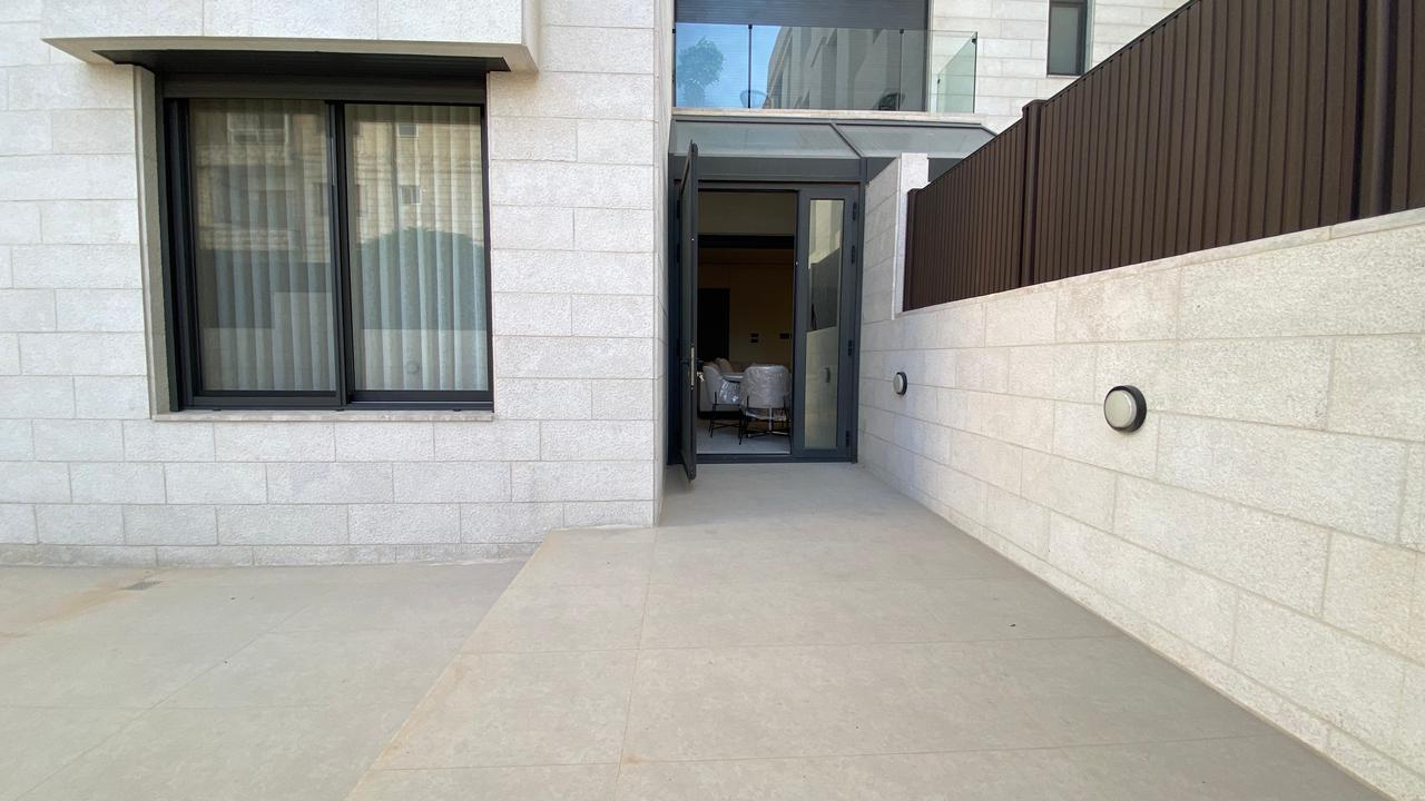 Open glass door into a modern apartment; beige stone walls, outdoor patio, and seating visible inside.