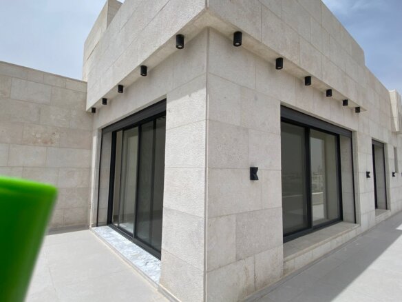Modern beige stone building with large black-framed glass doors/windows along a walkway; foreground shows a blurry green object partially covering the lower left corner.