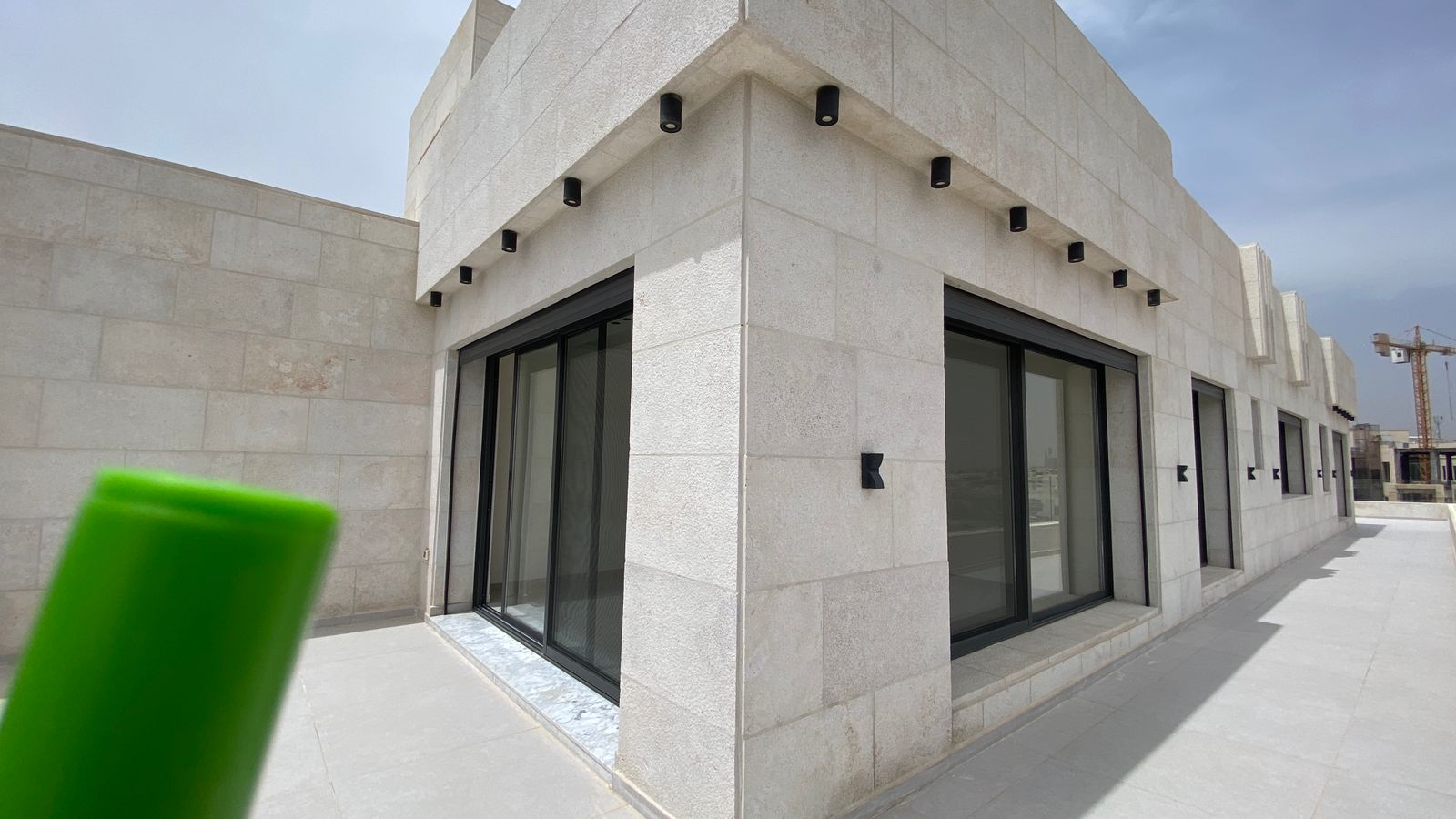b6ce51ff-bce7-4906-9ebc-b130449f0e8e Modern beige stone building with large black-framed glass doors/windows along a walkway; foreground shows a blurry green object partially covering the lower left corner.
