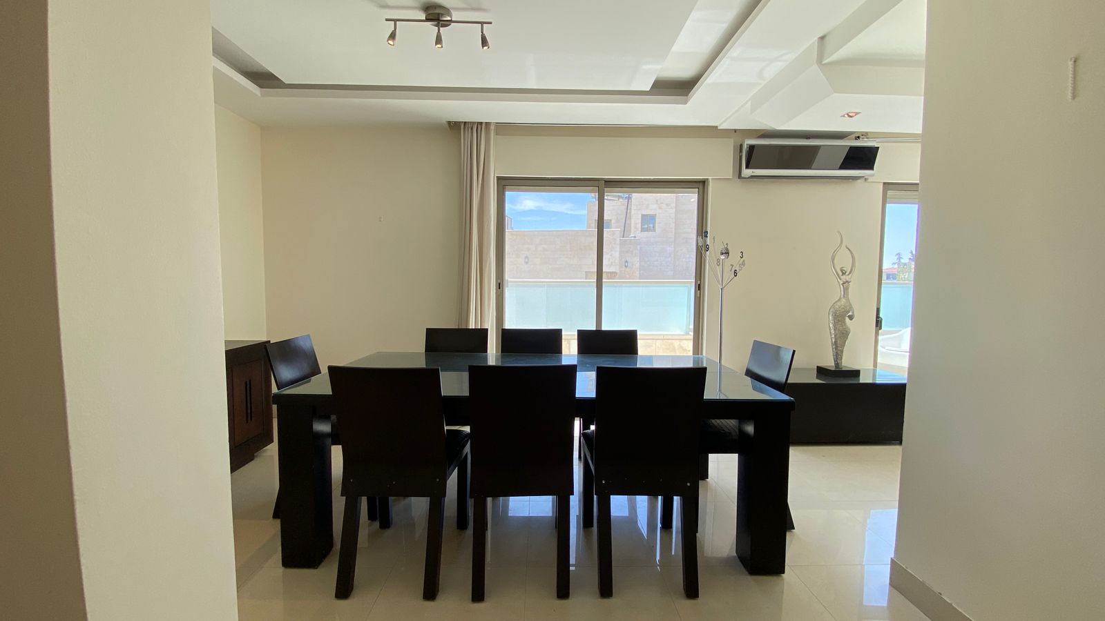 b738c36b-02b5-414c-bb1c-5c5b5401f474 Contemporary dining room with a glass-top table, dark wooden chairs, and a sliding glass door to a balcony letting in daylight.