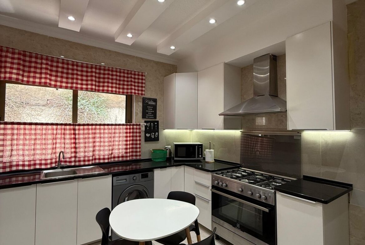 Modern kitchen with white cabinets, stainless steel oven/hood, black countertops, and red plaid window curtains; round white table with black chairs in foreground.