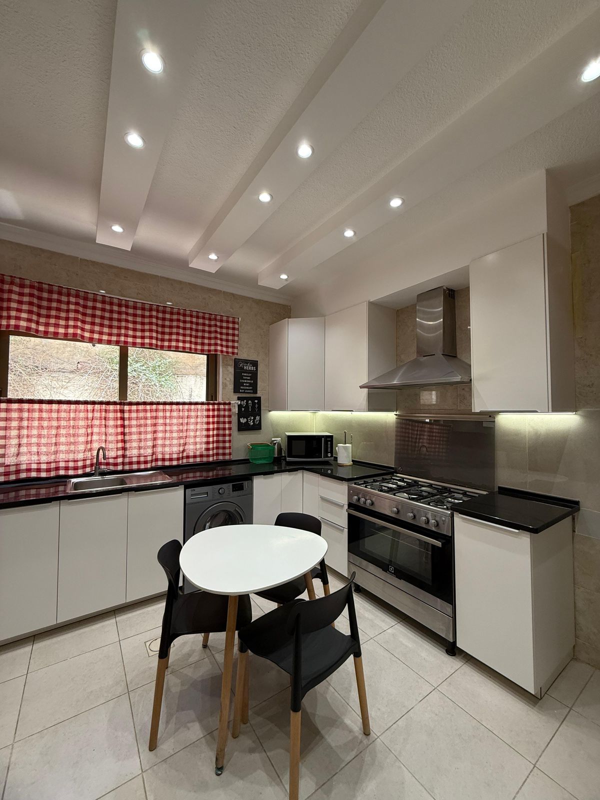 Modern kitchen with white cabinets, stainless steel oven/hood, black countertops, and red plaid window curtains; round white table with black chairs in foreground.