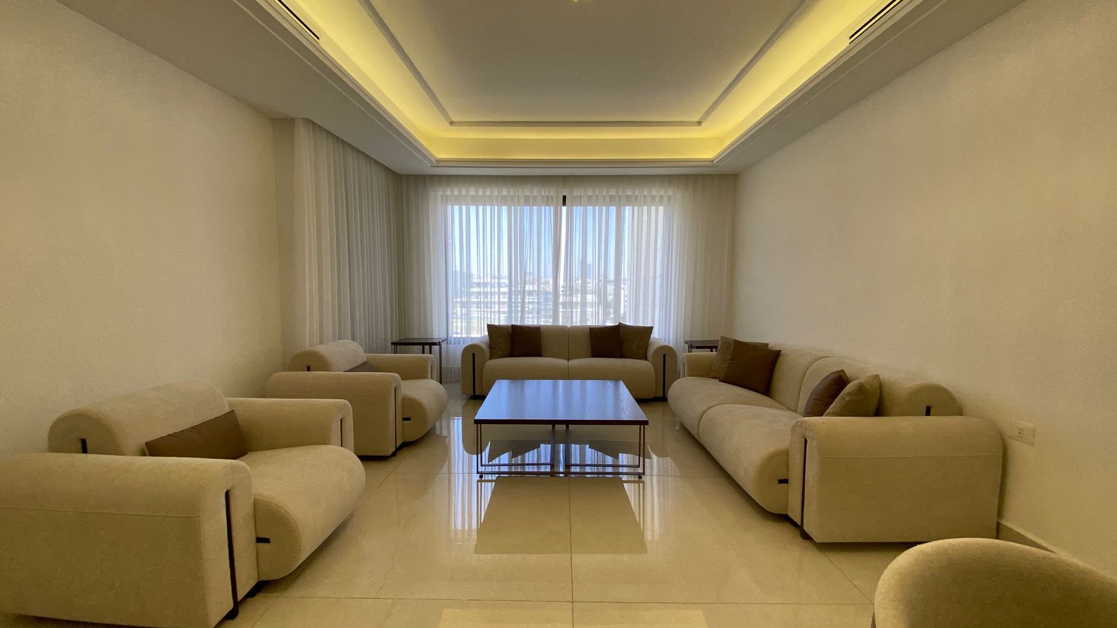 b830ff64-72e5-4b1b-b7d5-11bffb4d5a96 Neutral-toned living room with beige sofas around a central coffee table and sheer curtains over large windows