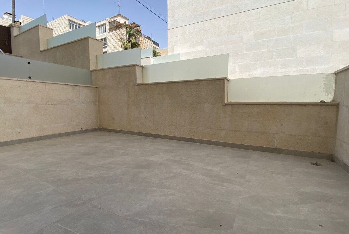 A rooftop terrace with beige stone walls, light gray tiled floor, and stepped frosted glass barriers along the edges under a blue sky.
