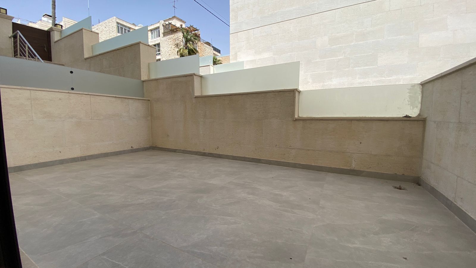 b88a5a86-e7d4-4be6-ae0f-a2d3c03f7ca5 A rooftop terrace with beige stone walls, light gray tiled floor, and stepped frosted glass barriers along the edges under a blue sky.