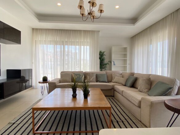 Bright open living room with a beige L-shaped sofa, striped rug, and large sheer curtains letting in daylight.