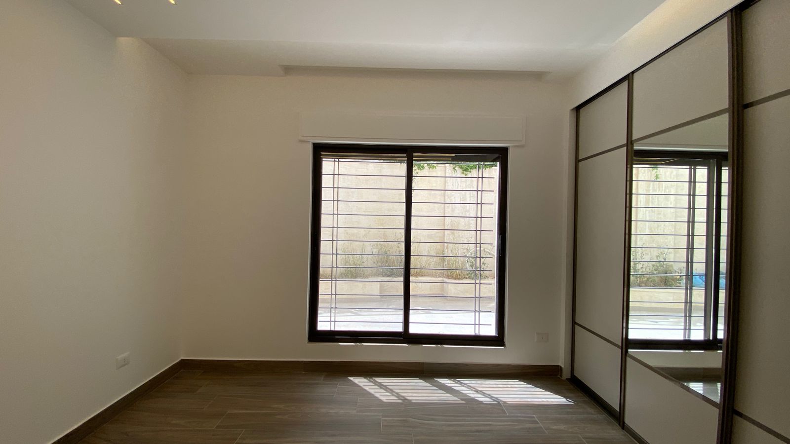 b97b6af4-8142-4fa4-99fc-ac2cc0591ca0 A small empty room with a large window barred by metal rails, light entering and casting shadows on wood-like flooring, right side shows a sliding closet with mirrored panels.