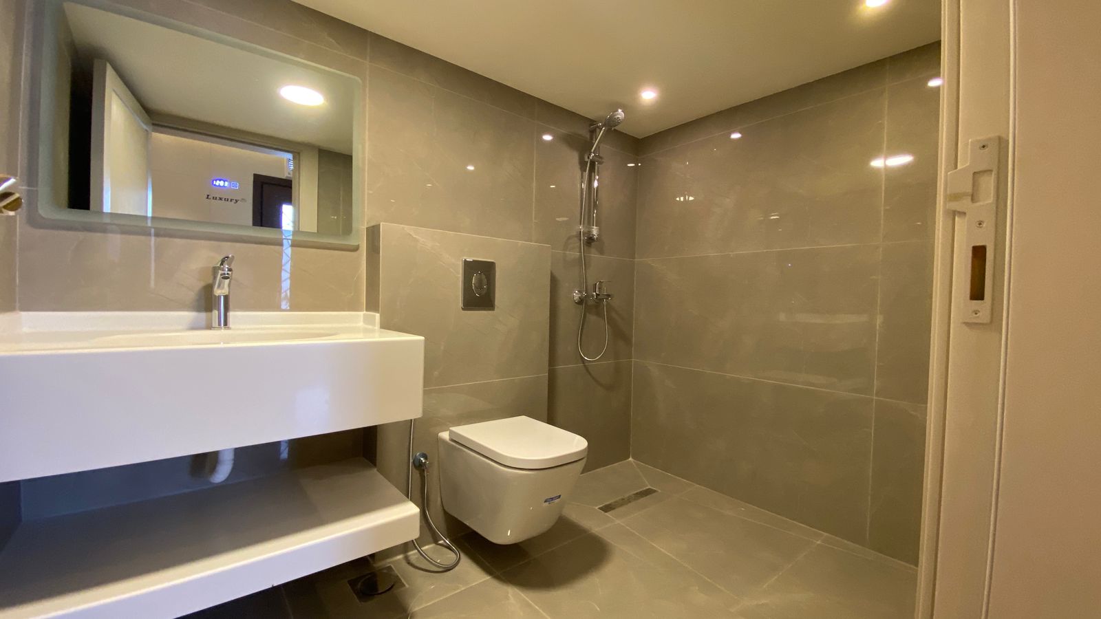 b9a10364-3317-408e-a357-3bc677ea4288 Modern hotel-style bathroom with gray tiled walls and floor, wall-mounted toilet, white sink, and a walk-in shower area