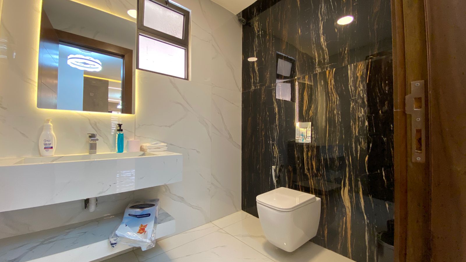 b9ae8bef-dd55-4b12-a63b-a2b0834d25ee Modern bathroom with white marble walls, a backlit mirror above a floating sink, and a white wall-mounted toilet near a black marble accent wall in a shower area.