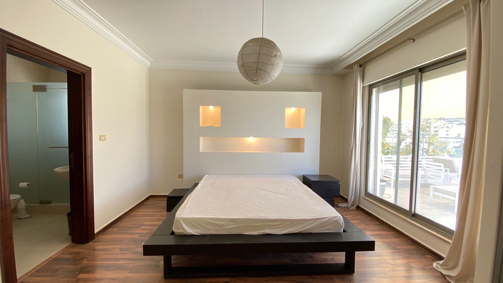 ba1f3129-e251-4cb9-a40b-f1d8a8c45429 Bright, minimalist bedroom with a low dark wood platform bed, white bedding, and a white backlit headboard featuring two square niches and a central light strip.
