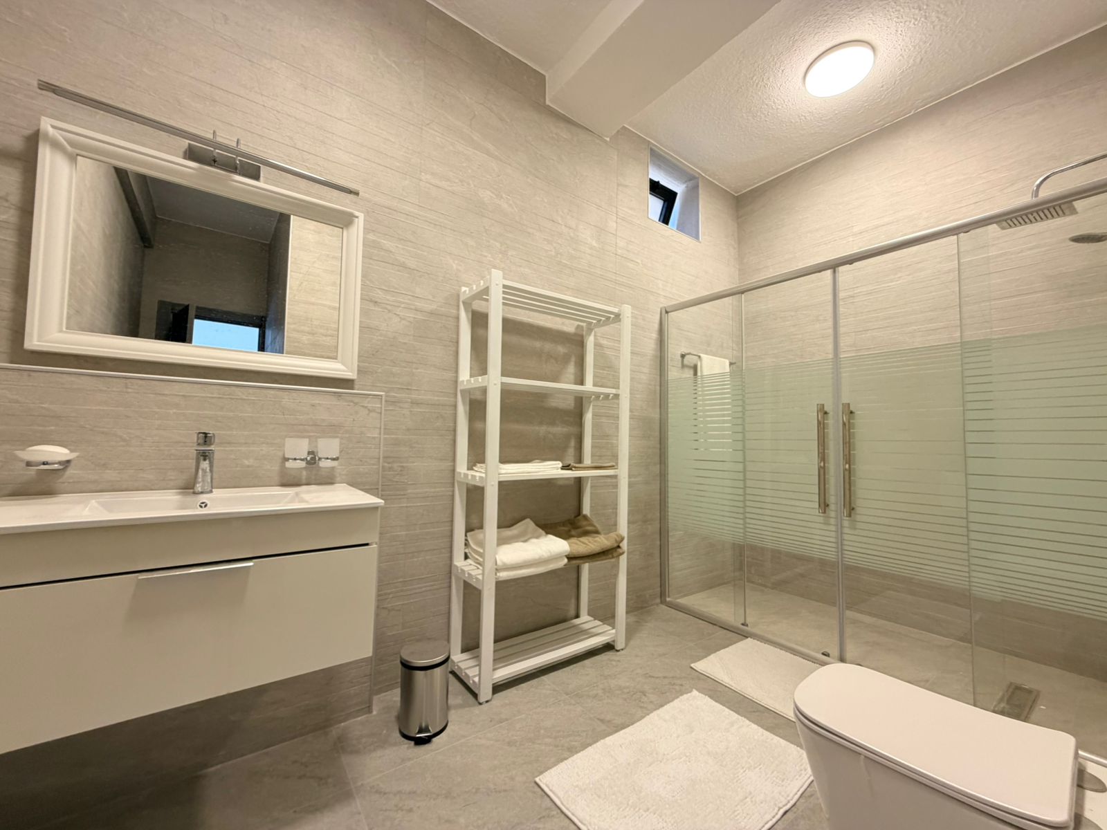 bab9765c-ebfd-4690-a2e2-a54d6c8b2a6e Modern bathroom with a white vanity, large framed mirror, and open shelving next to a glass shower enclosure.