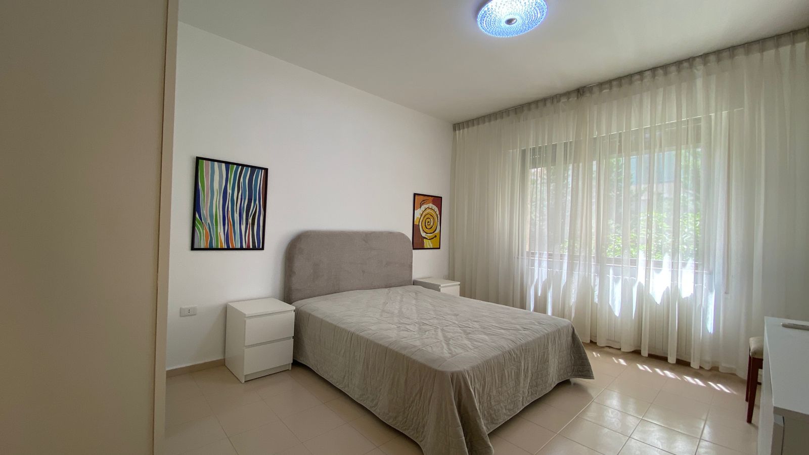 bad13a74-71fe-47c6-aae8-6842b91d8599 Bright, minimalist bedroom with a gray upholstered double bed, two white nightstands, and abstract wall art by a large window with sheer curtains