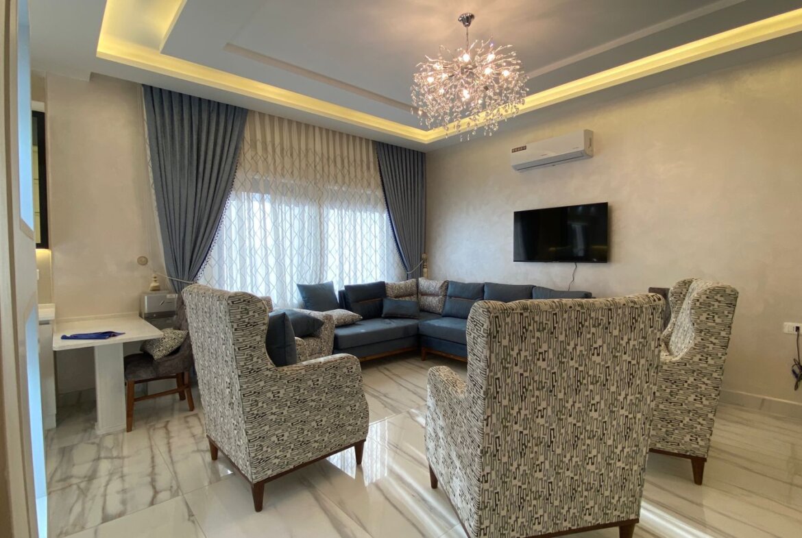 Living room with a blue sectional sofa and patterned armchairs around a marble floor, a crystal chandelier overhead.