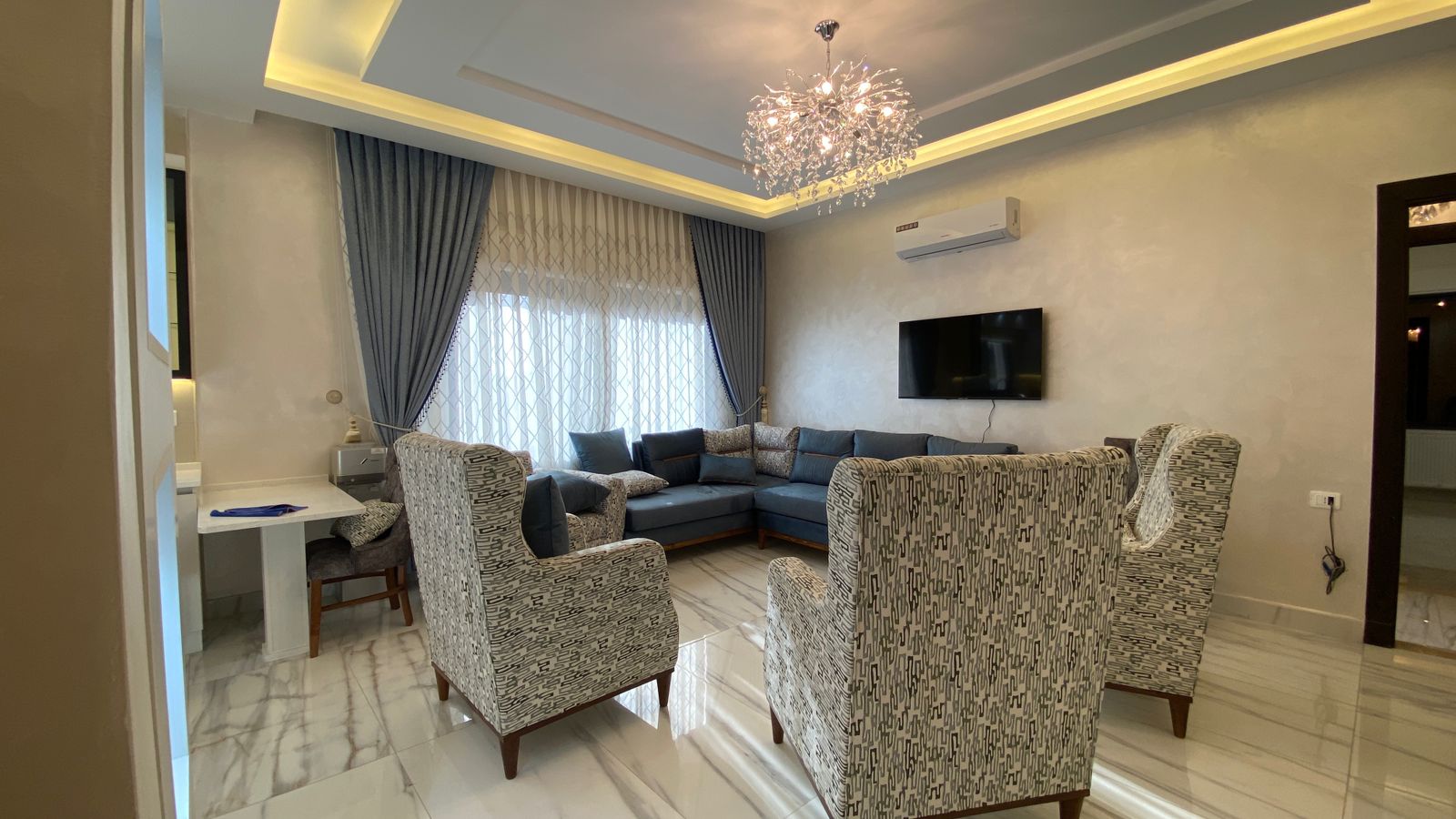 Living room with a blue sectional sofa and patterned armchairs around a marble floor, a crystal chandelier overhead.