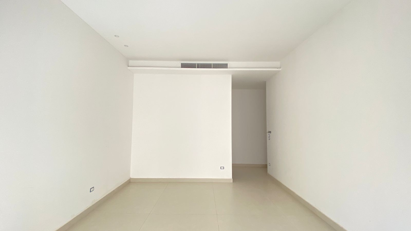 bc72019f-c7ec-4905-8fe5-6f0d4ca41ab6 Empty white room with a partial central wall, beige tiled floor, and a ceiling vent along the top; doorway at the far end.