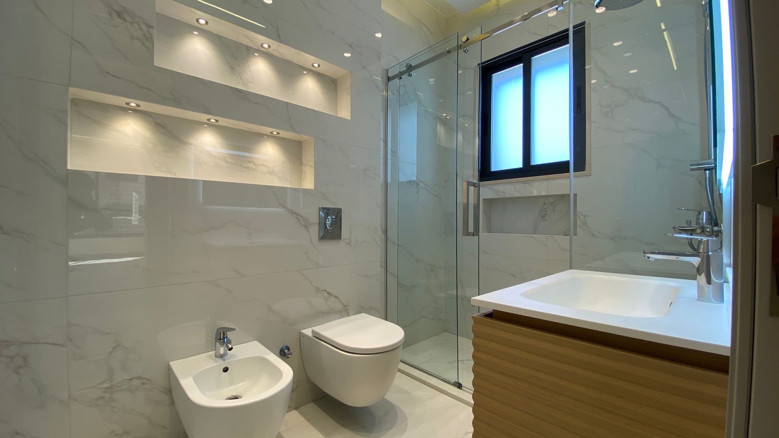 bca83a85-fcb7-43d3-8ae6-426a0ce37666 Modern marble bathroom with glass shower enclosure, wall-mounted toilet and bidet, and a wooden vanity with white sink.
