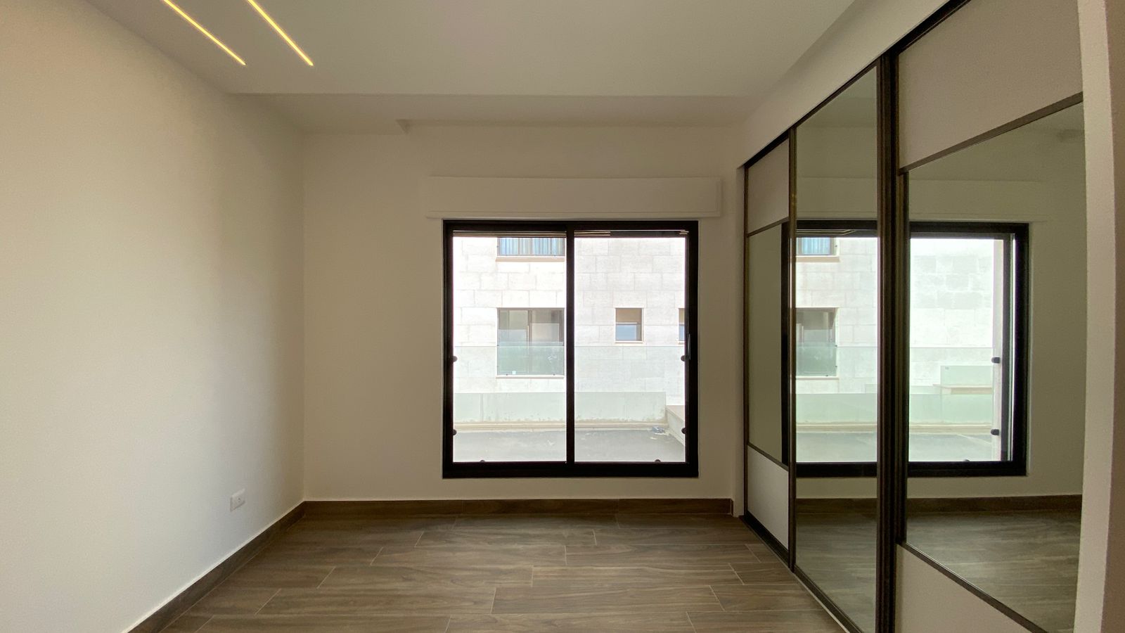 bcab13e2-a901-4ac5-962e-4ee74f8a356e Empty modern room with a large sliding glass window facing a pale building, and a mirrored wardrobe along the right wall.
