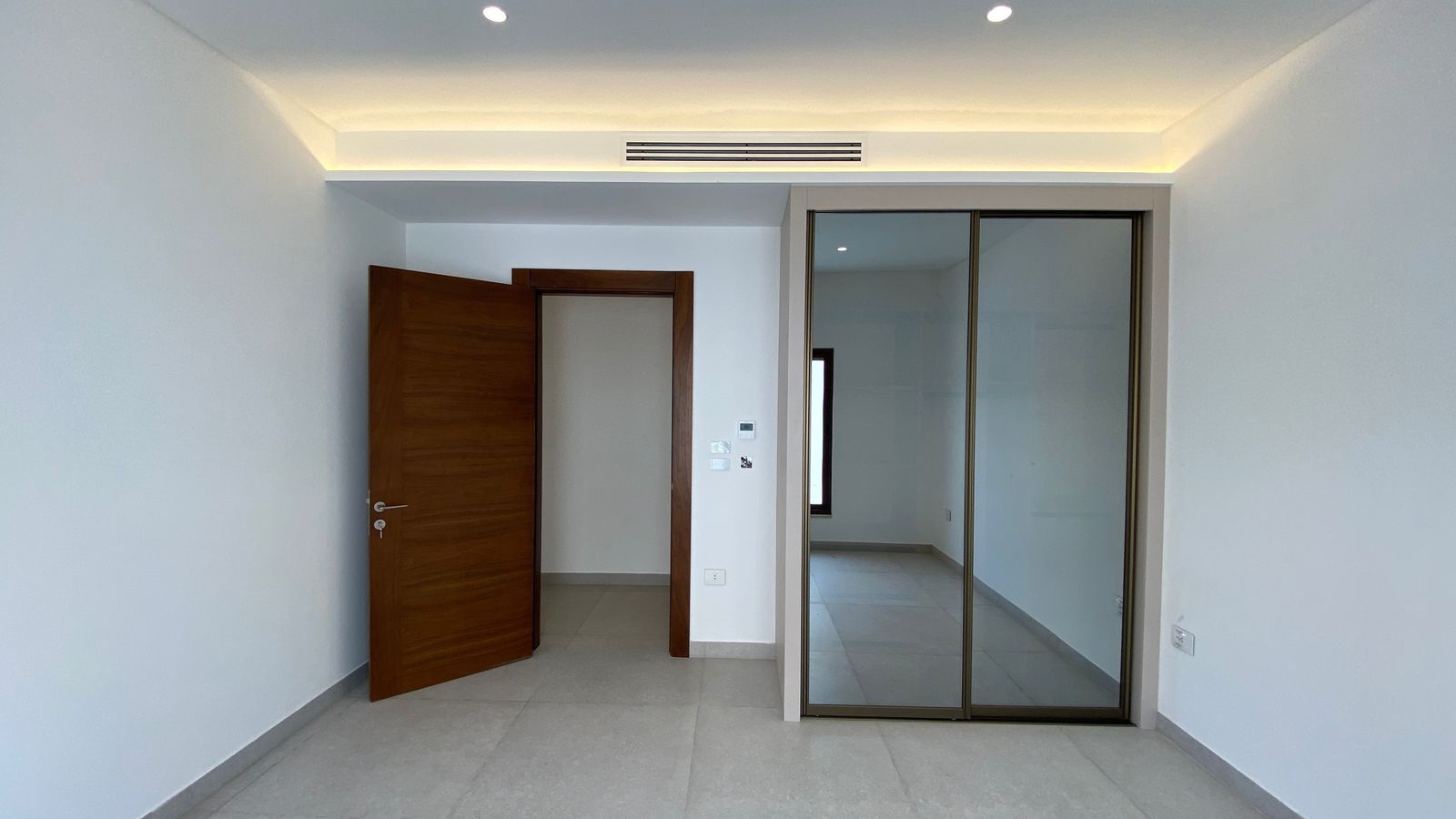 be6d55d6-af5a-4223-9438-a976326a1832 Empty, unfurnished room with beige tiled floor, open wooden door, and a glass sliding-cloak closet door; recessed ceiling lights above.