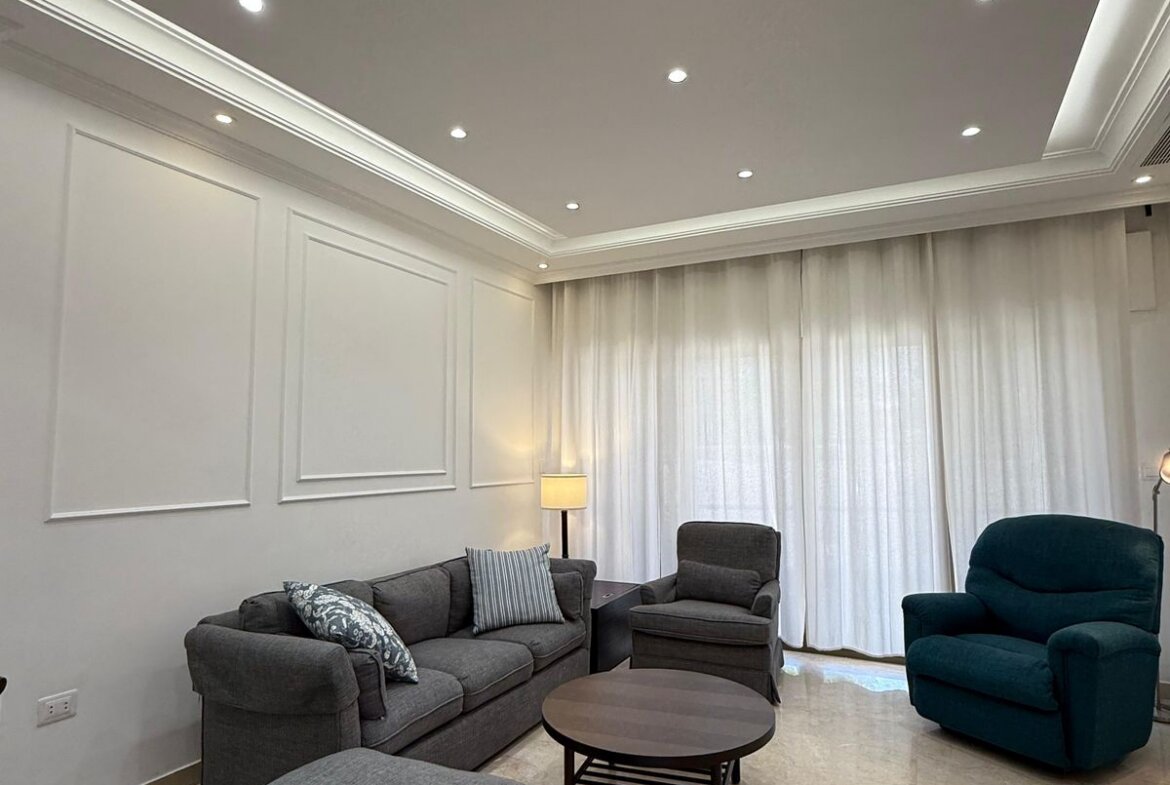 Modern living room with a gray sectional sofa, matching chairs, round coffee table, and white paneled walls under recessed ceiling lights.