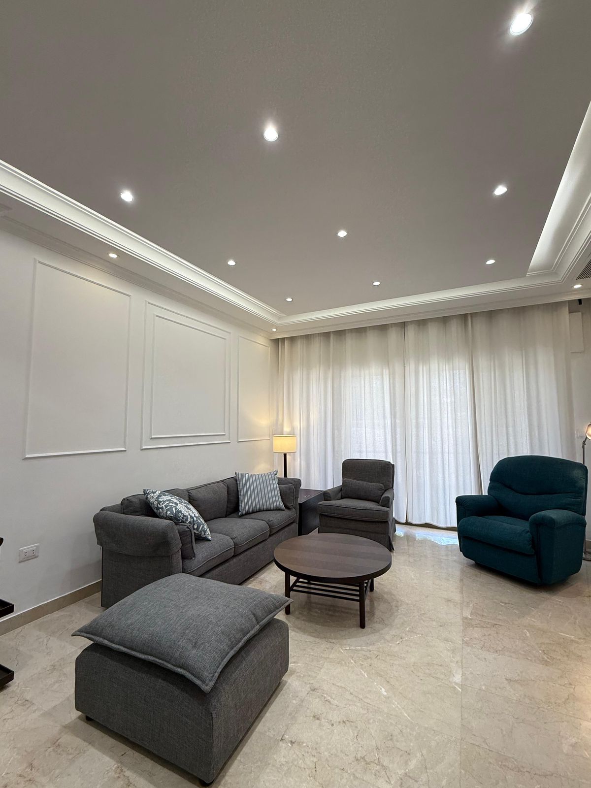 Modern living room with a gray sectional sofa, matching chairs, round coffee table, and white paneled walls under recessed ceiling lights.
