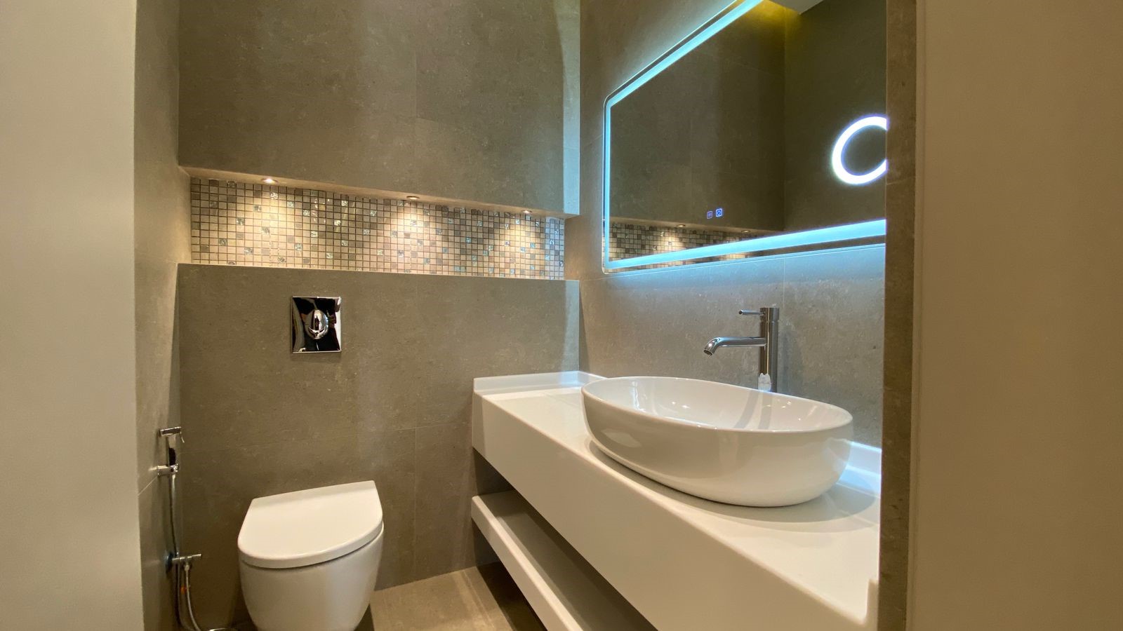 bf9e7fa4-07f9-4d9a-b242-9e1eeb68f6c1 Modern bathroom with a white wall-hung toilet, chrome bidet hose, and a white rectangular countertop supporting a round vessel sink under a backlit framed mirror. Mosaic tile strip above the sink adds detail to gray stone walls.