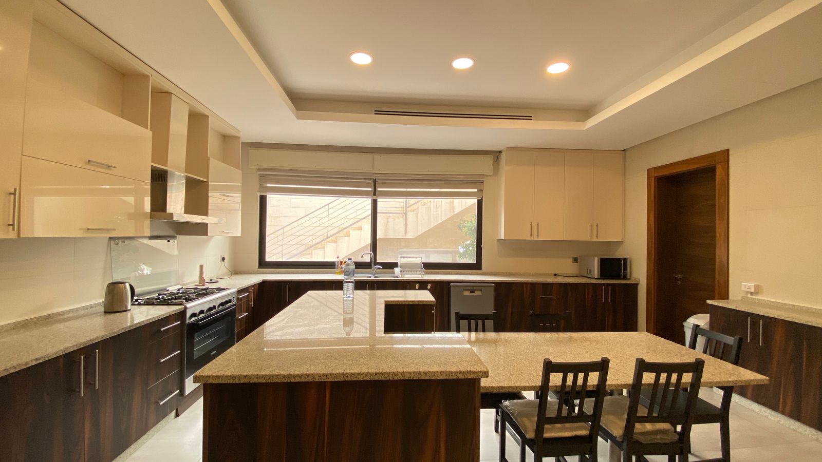 c009c83e-cbdf-4921-9c00-1fcb212363d1 Modern kitchen with a large granite island, dark wood base cabinets, white upper cabinets, and stainless appliances under a bright windowed wall.