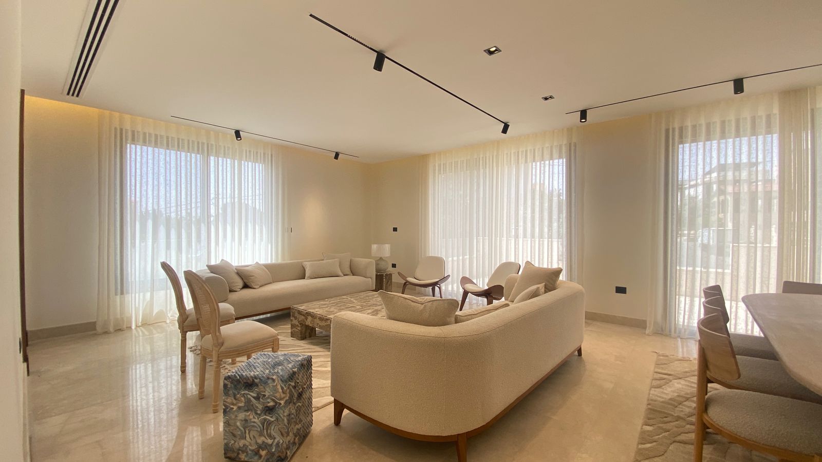 c0bc066c-3e65-47df-b759-9cff1a6ccd12 Bright, minimalist living room with white sofas, sheer curtains, and marble floor panels under track lighting.
