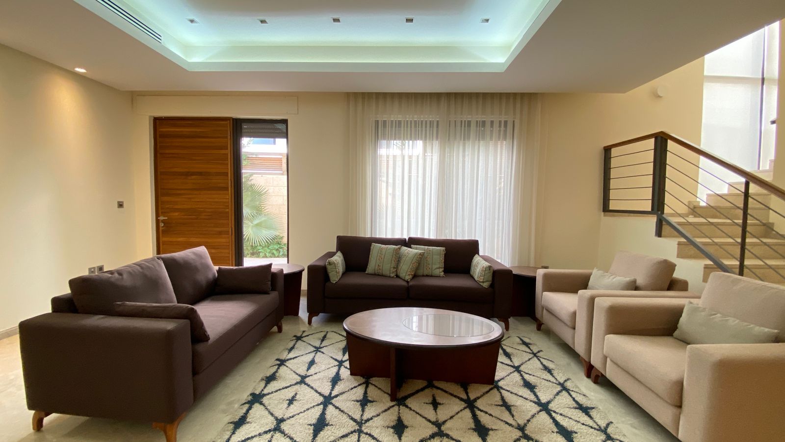 c0dc9c2f-1ded-4863-852a-85d0c3054393 Modern living room with brown sofas, beige armchairs, a round glass coffee table, and a geometric rug; staircase to the right.