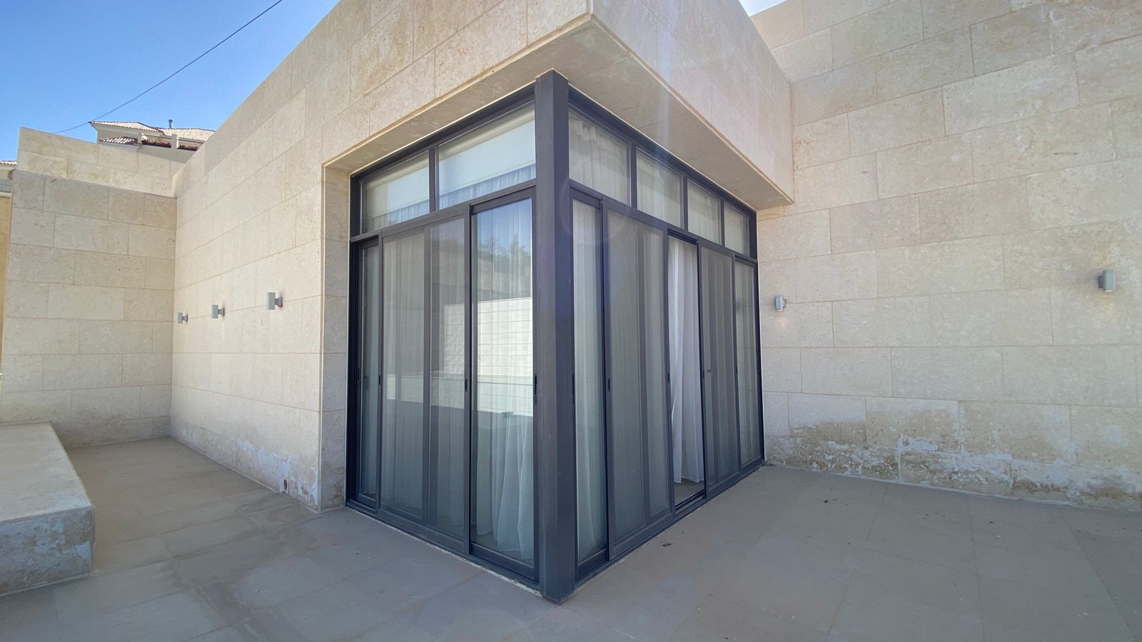 c1771991-f004-4ece-84df-73b53c0eb667 Corner of a modern beige stone building with large glass sliding doors framed in dark metal, leading to an outdoor patio.