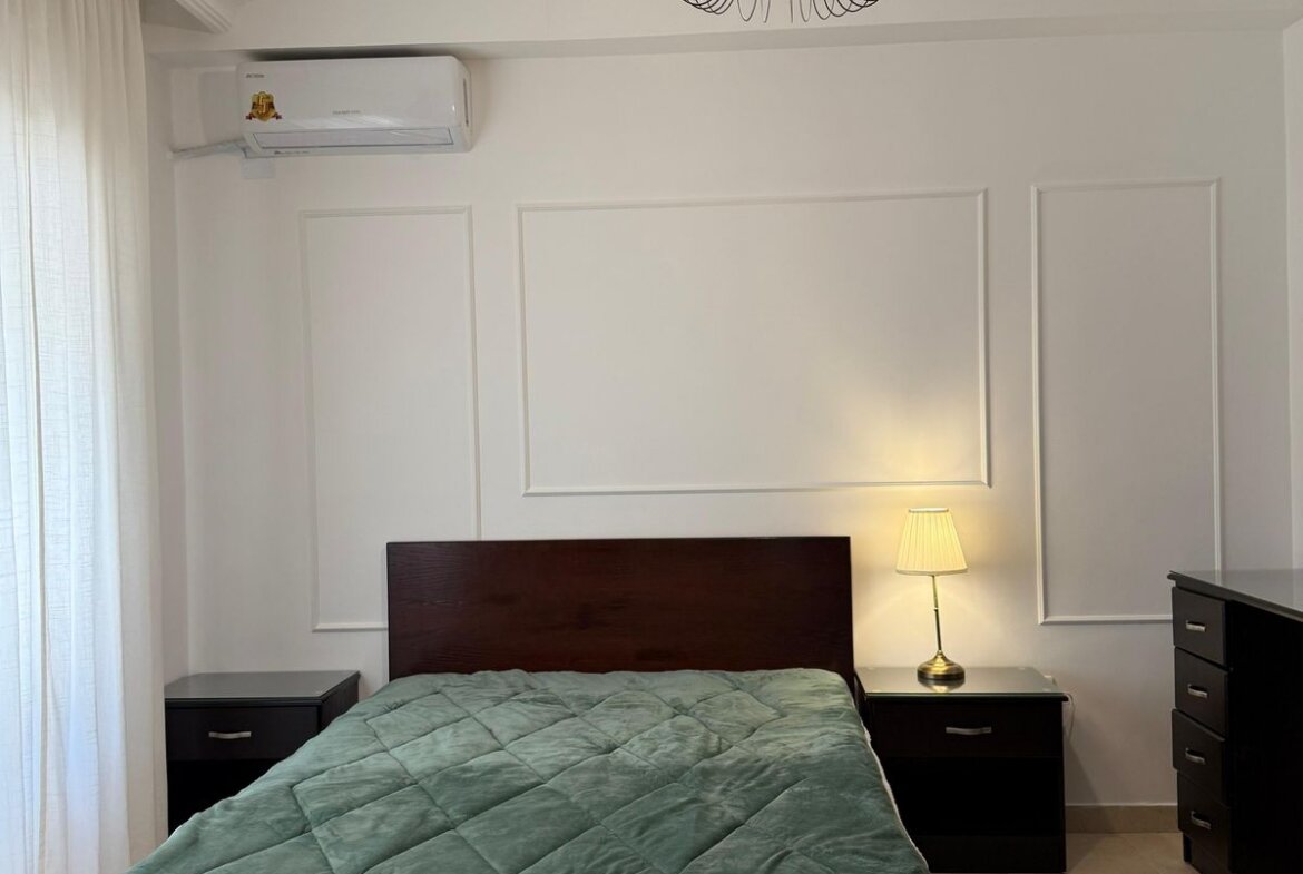 Bedroom with a dark wood bed, green quilt, matching nightstands, and a lamp on the right-side table; white paneled walls and a ceiling light fixture.