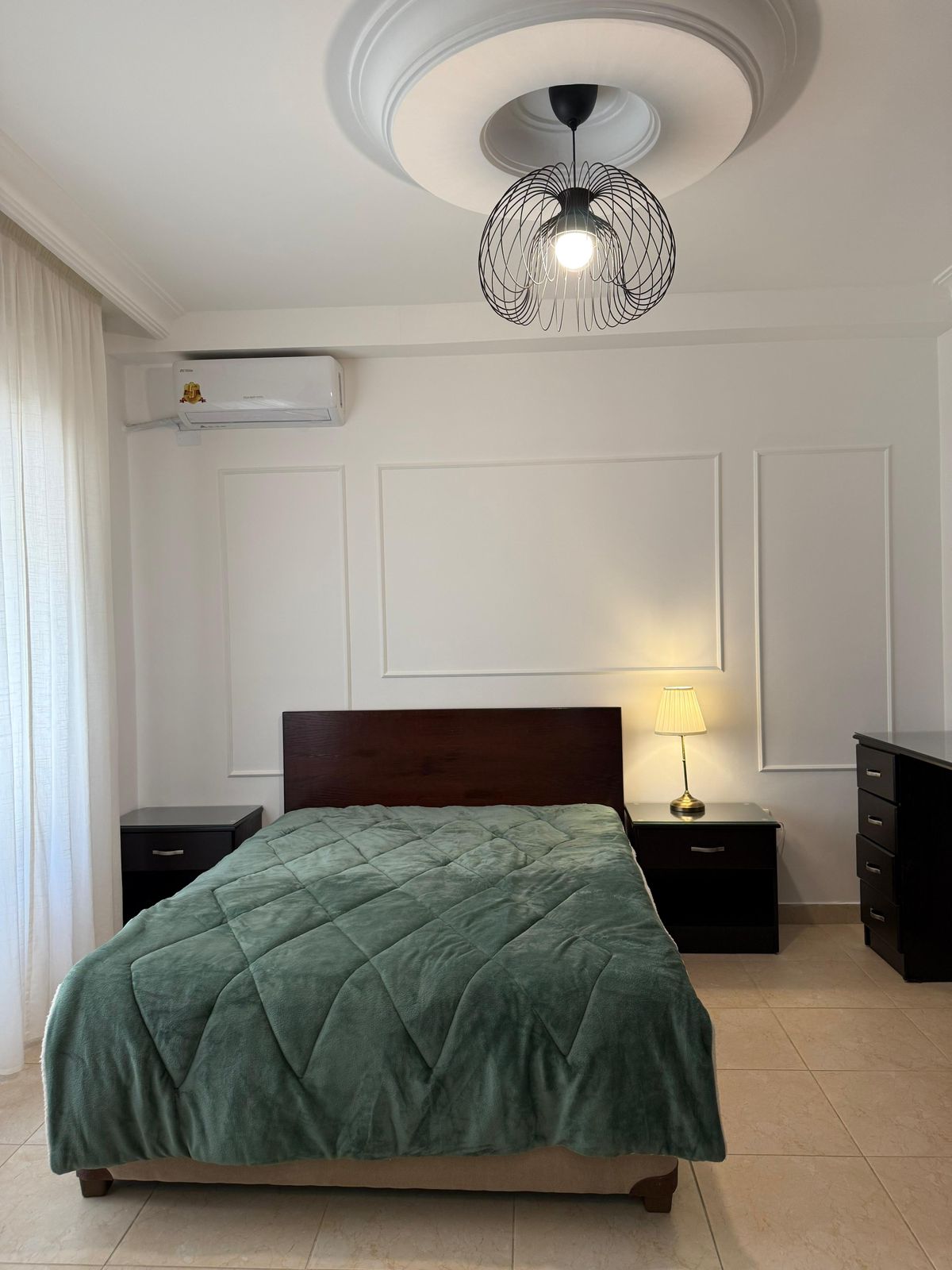 Bedroom with a dark wood bed, green quilt, matching nightstands, and a lamp on the right-side table; white paneled walls and a ceiling light fixture.