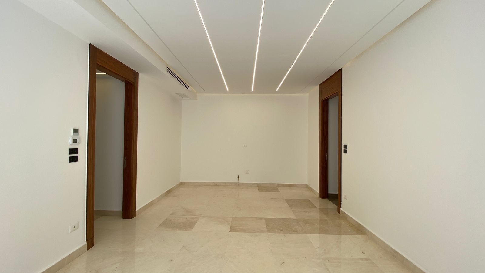 c213ebe5-eb80-45fe-8a28-ae16b6127f3a Empty rectangular room with white walls, beige marble floor, and wooden door frames; three parallel linear ceiling lights stretch across the ceiling.
