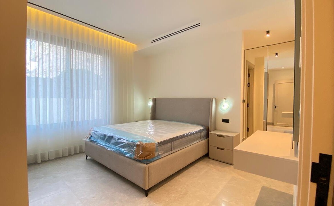 Modern hotel-style bedroom with a beige upholstered bed, two nightstands, circular wall lamps, sheer curtains, and a mirrored wardrobe. Mattress still wrapped in plastic in the foreground.]','Wide-angle view of a bright minimalist bedroom: large window with sheer curtains, an upholstered headboard bed, two small bedside tables, and a mirrored dresser.'],