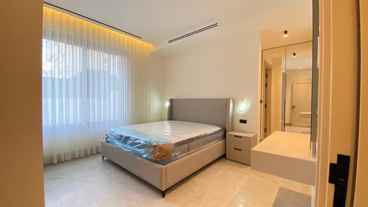 Modern hotel-style bedroom with a beige upholstered bed, two nightstands, circular wall lamps, sheer curtains, and a mirrored wardrobe. Mattress still wrapped in plastic in the foreground.]','Wide-angle view of a bright minimalist bedroom: large window with sheer curtains, an upholstered headboard bed, two small bedside tables, and a mirrored dresser.'],
